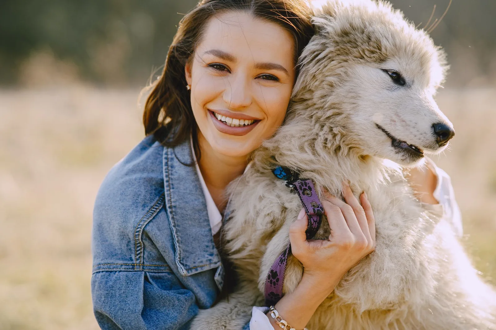 A smiling woman in a denim jacket hugging a happy, fluffy white dog outdoors.